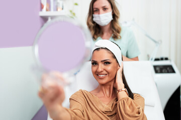 Beautiful and happy young woman sitting in medical chair and looking in the mirror. She is satisfied after successful beauty treatment with hyaluronic acid fillers or botulinum toxin injections.