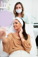 Beautiful and happy young woman sitting in medical chair and looking in the mirror. She is satisfied after successful beauty treatment with hyaluronic acid fillers or botulinum toxin injections.