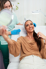 Beautiful and happy young woman sitting in medical chair and looking in the mirror. She is satisfied after successful beauty treatment with hyaluronic acid fillers or botulinum toxin injections.