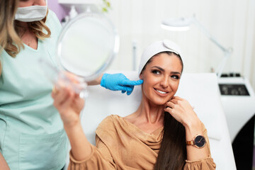 Beautician wearing blue gloves points at a patient's face, highlighting the results of a facial treatment in a beauty salon, while the woman looks happy and satisfied in the mirror