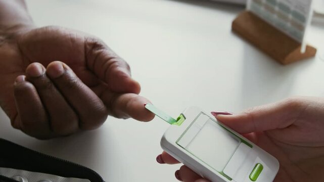 African American male patient holding hand with pricked finger on desk while female doctor with glucometer checking blood sugar level to find out or exclude diabetes