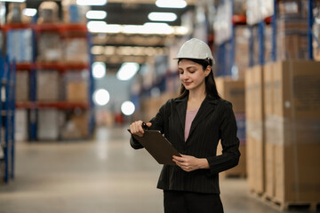 Professional woman working in warehouse. Wearing a white hard hat and holding clipboard. She checks the stock and goods in a large storage area.