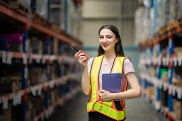 Smiling female warehouse worker in reflective vest holding a walkie-talkie and tablet. Standing in...