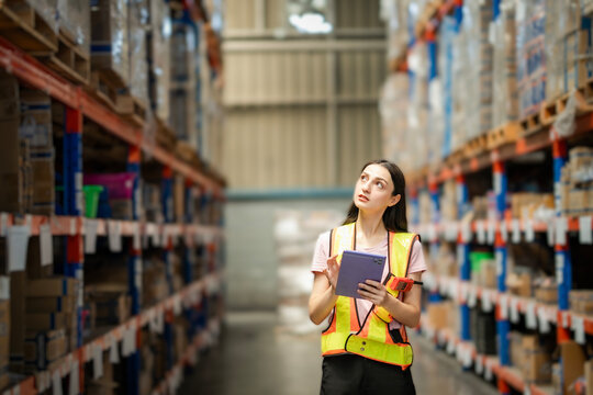 Young White female warehouse worker with safety vest holding tablet and barcode scanner. She is looking at stock while walking and checking between shelves. Inventory and logistics concept...