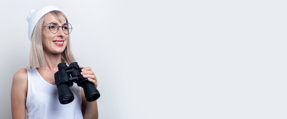 Cheerful young woman holding binoculars looks to the side on a light background. Banner