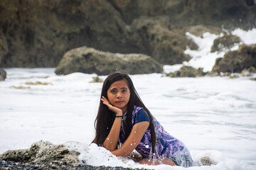 young woman with anxious surprised face sits on rocky sea beach wet by wave