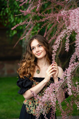 A beautiful young woman with long wavy hair in a floral vintage corset and black dress posing outdoors in a lush lilac garden. Springtime atmosphere, romantic and fairytale vibes. 