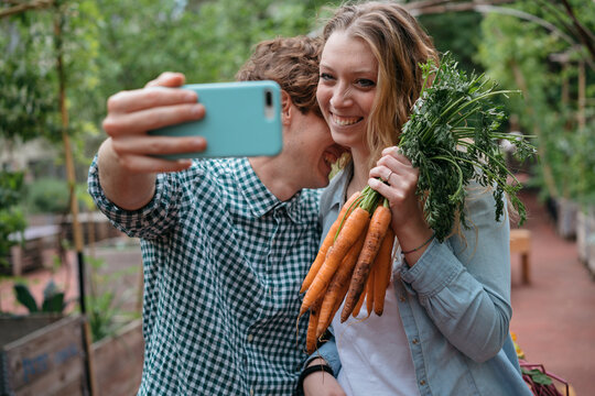 Smiling young man and woman take a selfie holding a bunch of fresh carrots. They are enjoying harvesting vegetables in their garden.