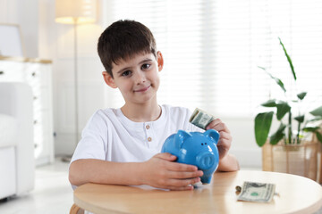 Boy putting pocket money into piggybank at table indoors