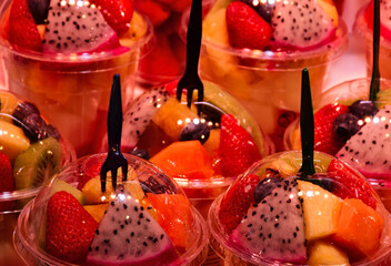 cups of colorful sliced fruits. transparent clear plastic cups. spherical caps. street vendor stand. closeup view. healthy eating and essential vitamins. freshness concept. peach, mango, dragon fruit