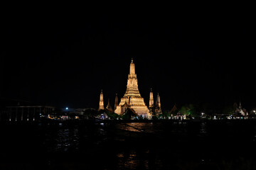 Naklejka premium Wat Arun, the Temple of Dawn, illuminated at night in Bangkok, Thailand. The golden lights highlight the intricate architecture, reflecting on the Chao Phraya River under a deep black sky.