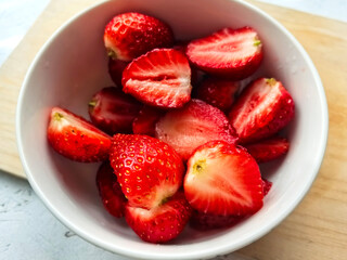 Sliced strawberries lie in a white plate against a wooden board and a white table. Macro photography of strawberries. Ripe red berries. Crops and fruits from the home garden