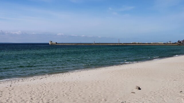 Scenic view of a quiet sandy beach and calm sea waves under a clear blue sky. A breakwater structure with buildings and a watchtower stretches along the horizon in the distance.
