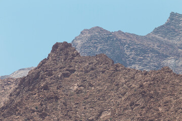 A rugged mountain landscape in Yazd, Iran, showcasing the arid, rocky terrain and layered geological formations under a clear blue sky.
