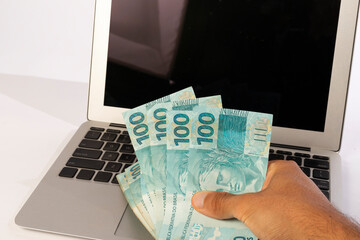 hands of a man holding Brazilian banknotes in front of a computer white background