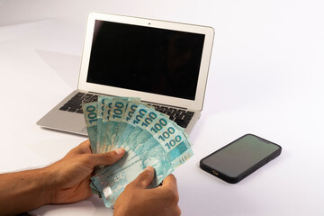 hands of a man holding Brazilian banknotes in front of a computer white background