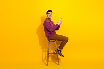 Young man wearing stylish casual clothing sitting on a stool against a yellow background holding a...