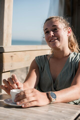Beautiful young woman enjoying an espresso served on a terrace with the ocean in the background.