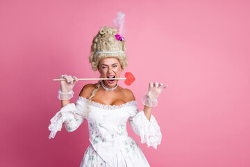 Elegant woman in baroque-inspired costume holding heart prop against pink backdrop, blending historic and playful styles