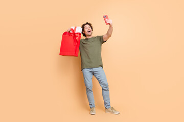 Excited young man taking a selfie while holding a red shopping bag against a beige background,...