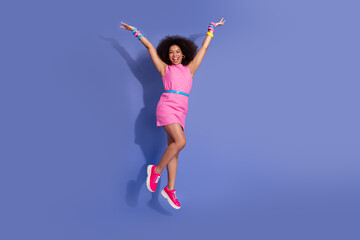Smiling young woman in a pink dress jumping joyfully against a purple background, expressing...