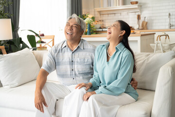 Asian senior couple laughing and smiling together on couch. Retired grandparent relaxing at home in the morning. Healthy elderly people and life insurance concept