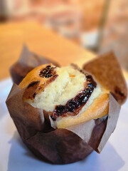 A close-up of a freshly baked muffin with chocolate swirls, wrapped in brown parchment paper, placed on a white plate in a warm, cozy cafe environment.
