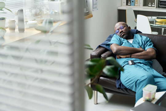 Tired medical professional sleeping in clinic break room, wearing scrubs, with medical equipment visible in background. Scene captures exhaustion of healthcare workers