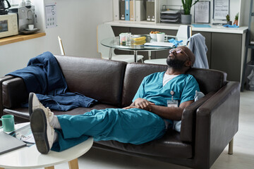 Medical professional wearing blue scrubs taking a break in hospital staff room with various office supplies and files visible in background