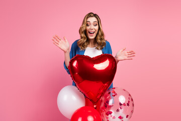 Joyful young woman in casual blue jacket posing with colorful balloons and a red heart-shaped...