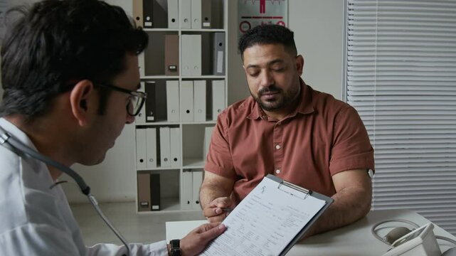 Male patient of medical center sitting in front of clinician and passing him card with personal information and diagnosis, doctor looking through data and consulting the man