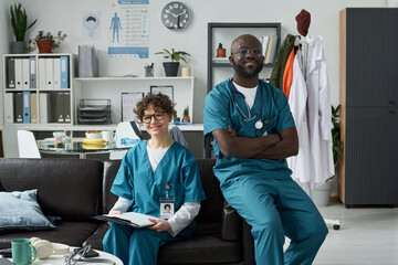 Portrait of two medical professionals sitting in office, one male and one female, both smiling and wearing scrubs, medical posters and equipment visible in background