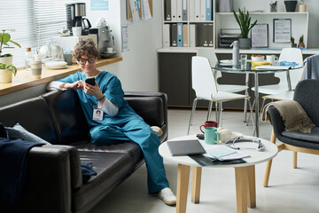 Female nurse taking break on couch in hospital break room while holding smartphone and drinking...
