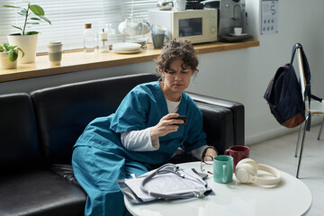 Healthcare worker wearing scrubs drinking coffee while sitting on couch in break room with medical...