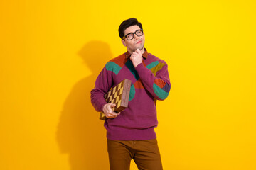 Young man in colorful sweater holding checkered game board, pondering decision against vibrant...