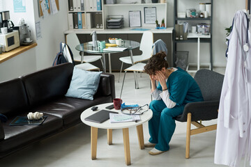 Healthcare worker sitting in office with hands on head, surrounded by medical equipment and files, displaying stress and fatigue. Office includes couch, table, and shelving unit
