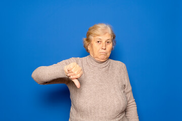 Caucasian woman in her 70s wearing a dark beige sweater with her thumb down in a sign of disgust and disapproval, isolated on blue studio background.