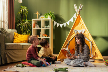Girl reading a story to two boys in playroom with teepee