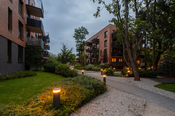 Illuminated pathway in landscaped courtyard of modern residential complex at dusk. Bollard lights create warm ambiance among lawns, shrubs, trees, near apartment buildings. A peaceful community space.