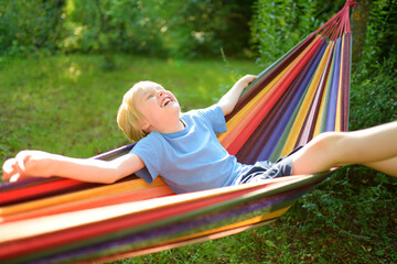 A cute blonde white preteen boy is having fun in the summer in a colorful hammock in the backyard or on the outdoor playground. A child is resting and swinging in a hammock.
