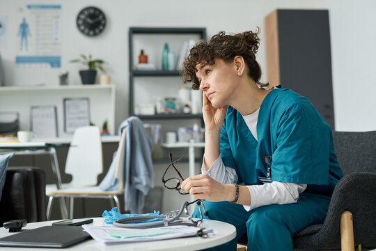 Healthcare professional in scrubs sitting at desk in hospital office, holding glasses and resting head on hand, appearing deep in thought about work commitments