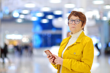 Portrait of elderly woman during shopping at mall. Red-haired senior lady using smartphone