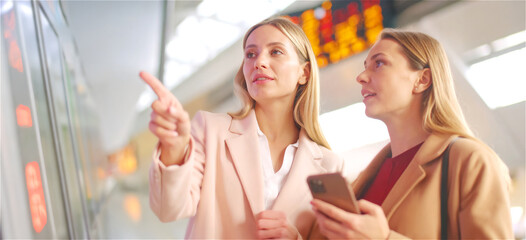 Two women at a train station comparing the travel data between smartphone and info display