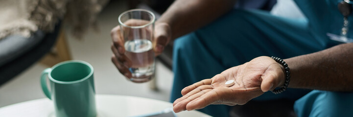Close-up of person holding white pill in one hand and glass of water in the other, with focus on hands and pill. Background shows some elements of the room