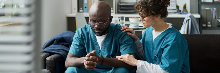 Nurse comforting a distressed healthcare worker sitting in a break room. Medical uniforms and professional workspace suggesting a healthcare setting