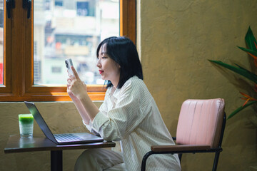 Young woman engaging with smartphone while seated at a table in a cozy cafe with a laptop and...