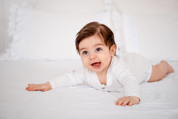 Fototapeta premium a baby on a white bed at home, a place for text, a cute smiling little child getting ready for bed, a portrait of a little boy child