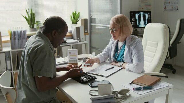 African American man pricking his finger with pen while female clinician with glucometer monitoring his blood sugar level and showing him data on device screen during medical consultation