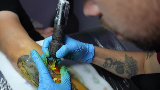 Close-up view of a tattoo artist in blue gloves creating a floral tattoo on a clients forearm. The tattoo machine is in motion as the artist outlines the design. Studio environment