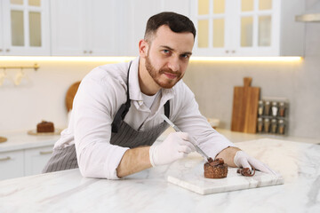 Confectioner decorating delicious dessert with piece of chocolate at white marble table in kitchen